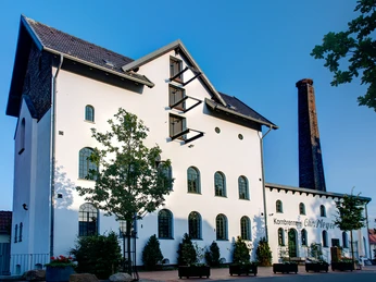 Alte Brennerei Hille Ehemalige Brennerei mit weißer Fassade, Ziegeldach und hohem Schornstein vor blauem Himmel.