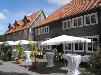 Älteres Fachwerkhaus mit roten Ziegeldächern, davor geschmückte Terrassen mit weißen Sonnenschirmen.