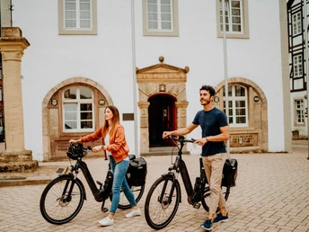 Zwei Personen schieben ihre E-Bikes über den gepflasterten Marktplatz vor historischen Gebäuden.