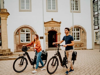 Zwei Personen schieben ihre E-Bikes über den gepflasterten Marktplatz vor historischen Gebäuden.
