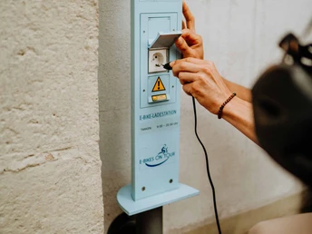 Teutoburger Wald-Brakel-Tourist Info ┬® Teutoburger Wald Tourismus (1).jpg Person lädt ein E-Bike an einer blauen Ladestation mit dem Logo "VORWEG FAHREN" an einer Wand auf.