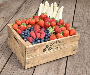 Spargelhof Winkelmann Eine Holzkiste auf Holzboden gefüllt mit frischem Obst und weißem Spargel, darunter Erdbeeren, Himbeeren und Blaubeeren.