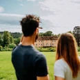 Ein Mann und eine Frau blicken auf Schloss Rheder, umgeben von üppigen grünen Wiesen und Wäldern.