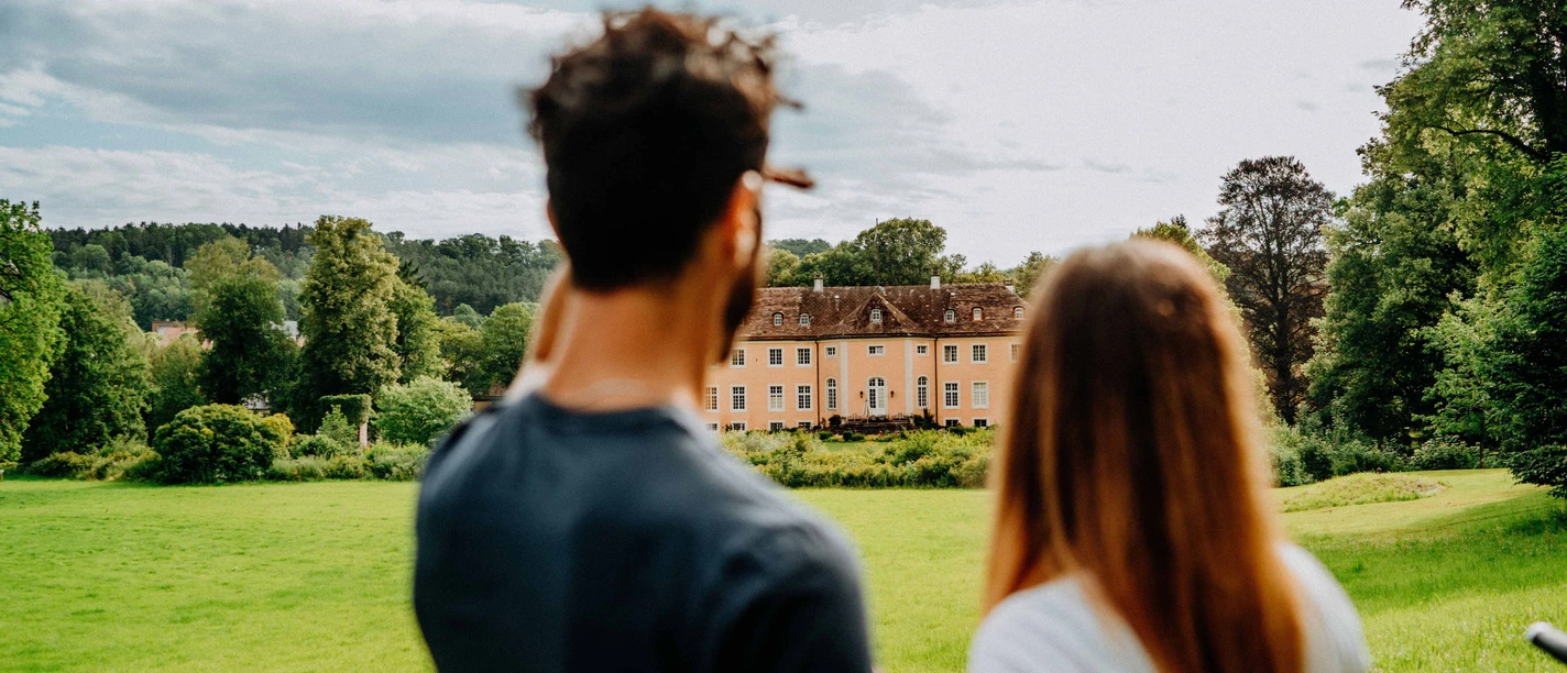 Blick auf Schloss Rheder - Brakel Ein Mann und eine Frau blicken auf Schloss Rheder, umgeben von üppigen grünen Wiesen und Wäldern.