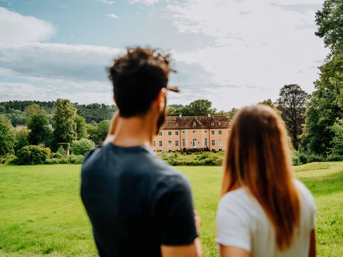 Blick auf Schloss Rheder - Brakel Ein Mann und eine Frau blicken auf Schloss Rheder, umgeben von üppigen grünen Wiesen und Wäldern.