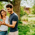 Zwei Menschen stehen lächelnd auf einer Wiese und betrachten ein Smartphone, ein Schloss ist im Hintergrund.