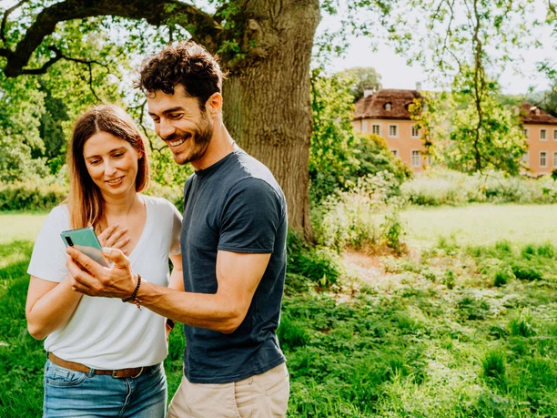 Zwei Menschen stehen lächelnd auf einer Wiese und betrachten ein Smartphone, ein Schloss ist im Hintergrund.