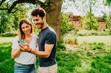 Teutoburger Wald-Brakel-Schloss Rheder Zwei Menschen stehen lächelnd auf einer Wiese und betrachten ein Smartphone, ein Schloss ist im Hintergrund.