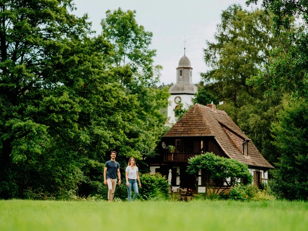 Zwei Personen spazieren vor einem historischen Fachwerkhaus mit Turm, umgeben von üppigem Grün.