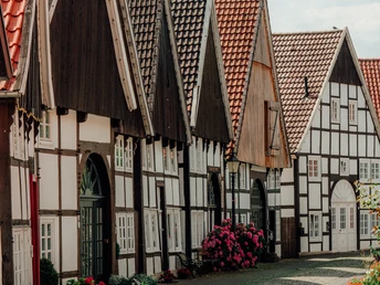 Großer_Wall_Rheda.jpg Eine malerische, gepflasterte Gasse mit traditionellen Fachwerkhäusern und blühenden Sträuchern.