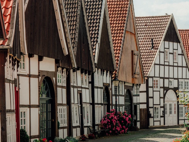 Großer_Wall_Rheda.jpg Eine malerische, gepflasterte Gasse mit traditionellen Fachwerkhäusern und blühenden Sträuchern.