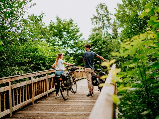 Zwei Personen schieben Fahrräder über eine hölzerne Brücke inmitten grüner, dichter Vegetation.