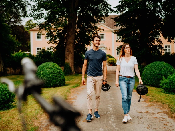 Ein Paar spaziert vor Schloss Rheder, umgeben von gepflegten Hecken und üppigem Grün.