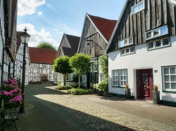 Großer Wall Rheda Fachwerkhäuser in einer gepflasterten Gasse mit blühenden Pflanzen und strahlendem Sonnenschein.