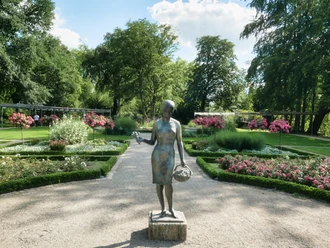 Skulptur einer Frau mit Korb in einem gepflegten Garten, umgeben von blühenden Blumenbeeten.
