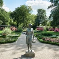 Rosengarten Rheda Skulptur einer Frau mit Korb in einem gepflegten Garten, umgeben von blühenden Blumenbeeten.