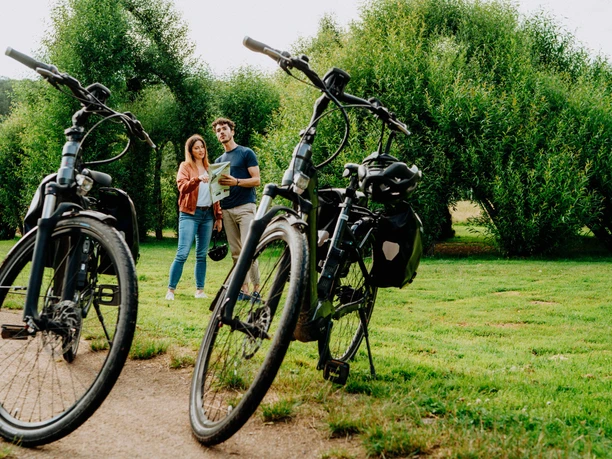 Zwei Personen stehen in einem grünen Park neben zwei Fahrrädern und studieren eine Landkarte.