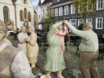 Alltagsmenschen Tänzerfiguren aus Stein in bunten Kleidern auf einem gepflasterten Platz vor historischen Gebäuden.