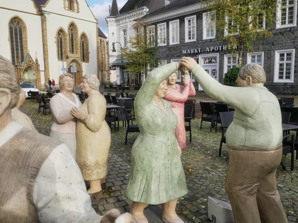 Alltagsmenschen Tänzerfiguren aus Stein in bunten Kleidern auf einem gepflasterten Platz vor historischen Gebäuden.