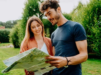 Ein Paar studiert stehend eine Landkarte in einem grünen Park mit Bäumen im Hintergrund.