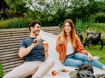 Weidenpalais Rheder Ein junges Paar sitzt auf einer Bank im Grünen, isst Obst und genießt einen entspannten Moment.