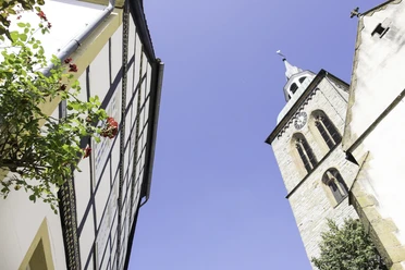 Fachwerkhaus, rote Blüten und Kirche mit Uhrturm, blauer Himmel im Hintergrund.