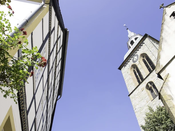 Fachwerkhaus, rote Blüten und Kirche mit Uhrturm, blauer Himmel im Hintergrund.