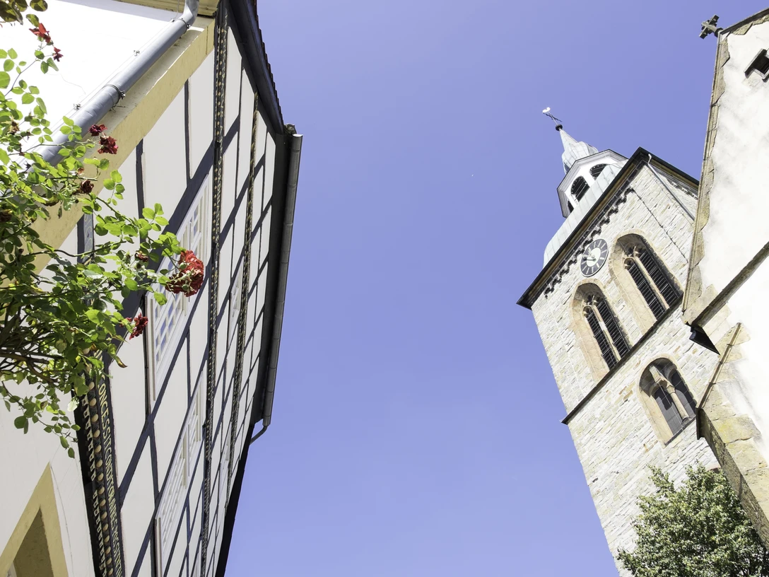 Giebel des Historischen Rathauses Wiedenbrück Fachwerkhaus, rote Blüten und Kirche mit Uhrturm, blauer Himmel im Hintergrund.