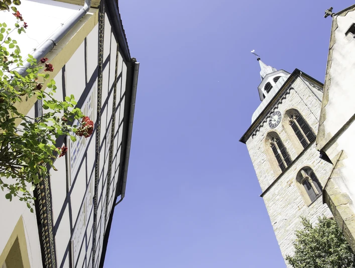 Giebel des Historischen Rathauses Wiedenbrück Fachwerkhaus, rote Blüten und Kirche mit Uhrturm, blauer Himmel im Hintergrund.