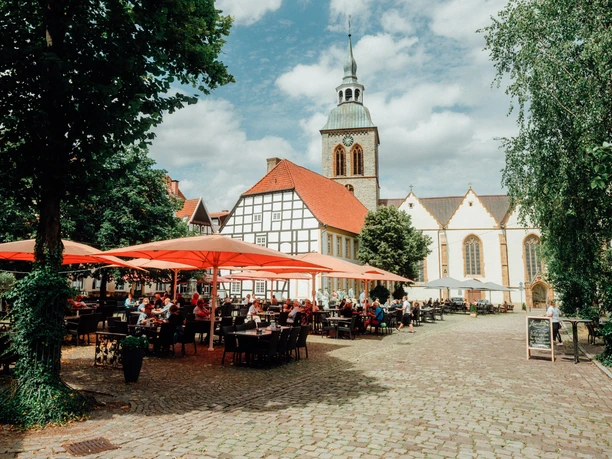 Historischer Platz mit Kirche, Fachwerkhäusern und einem Café im Freien unter roten Sonnenschirmen.
