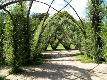 Ein kunstvoll gewölbtes Bauwerk aus geflochtenen Weiden, umgeben von üppigem Grün im offenen Freiraum.