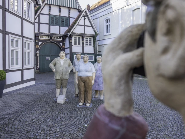 Alltagsmenschen Skulpturengruppe aus älteren Personen vor Fachwerkhäusern, im Vordergrund eine weitere Statue mit Kamera.