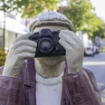 Skulptur eines Fotografen mit Kamera im Gesicht, in einer belebten, baumgesäumten Straße.