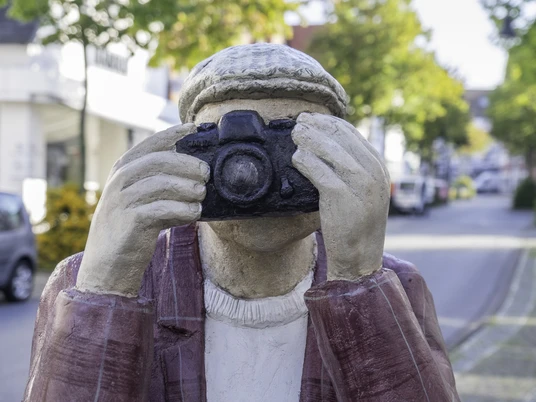 Skulptur eines Fotografen mit Kamera im Gesicht, in einer belebten, baumgesäumten Straße.