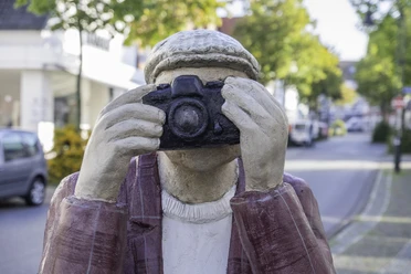 Alltagsmenschen in Wiedenbrück Skulptur eines Fotografen mit Kamera im Gesicht, in einer belebten, baumgesäumten Straße.