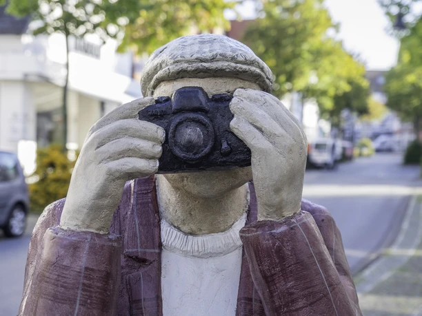 Alltagsmenschen in Wiedenbrück Skulptur eines Fotografen mit Kamera im Gesicht, in einer belebten, baumgesäumten Straße.
