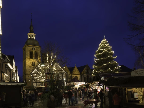 Historische Kirche mit beleuchtetem Weihnachtsmarkt am Abend, Besucher genießen die festliche Stimmung.