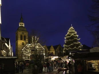 Christkindlmarkt Wiedenbrück.jpg Historische Kirche mit beleuchtetem Weihnachtsmarkt am Abend, Besucher genießen die festliche Stimmung.