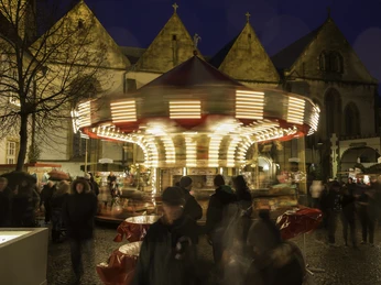 14-12 Christkindlmarkt Wiedenbrück 4585.jpg Ein beleuchtetes Karussell dreht sich im Abendlicht, umgeben von historischen Gebäuden und Menschen.