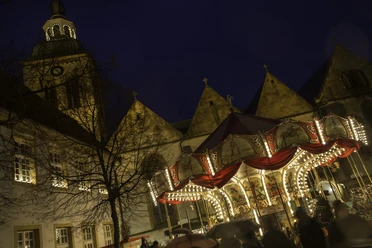 14-12 Christkindlmarkt Wiedenbrück 4589.jpg Historischer Weihnachtsmarkt bei Nacht mit Karussell und erleuchteten Gebäuden vor einem Kirchturm.
