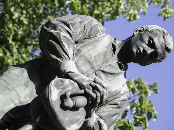 Bronzestatue eines Mannes in Arbeitskleidung mit historischem Werkzeug vor grünem Laub und blauem Himmel.