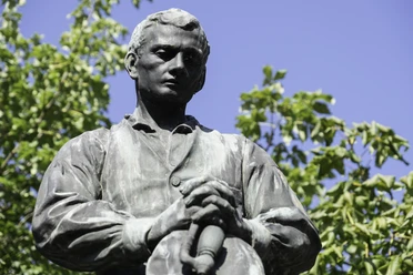 Bronzestatue von Adalbert Stifter in einem Garten, umgeben von grünen Bäumen unter klarem Himmel.
