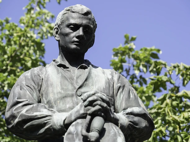 Bronzestatue von Adalbert Stifter in einem Garten, umgeben von grünen Bäumen unter klarem Himmel.