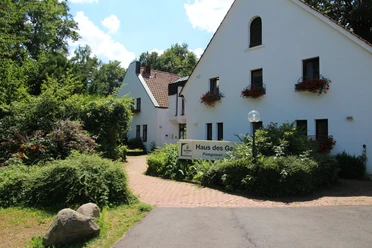 Weißes Gebäude mit begrünten Fassaden und blühenden Fensterkästen, umgeben von üppiger Natur.