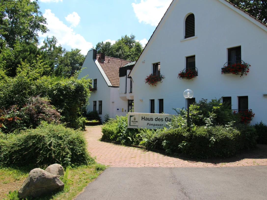 Haus des Gastes.JPG Weißes Gebäude mit begrünten Fassaden und blühenden Fensterkästen, umgeben von üppiger Natur.