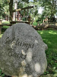 Großer Stein mit Inschrift „Schlumps Backs“, umgeben von Bäumen und einem traditionellen Fachwerkhaus.