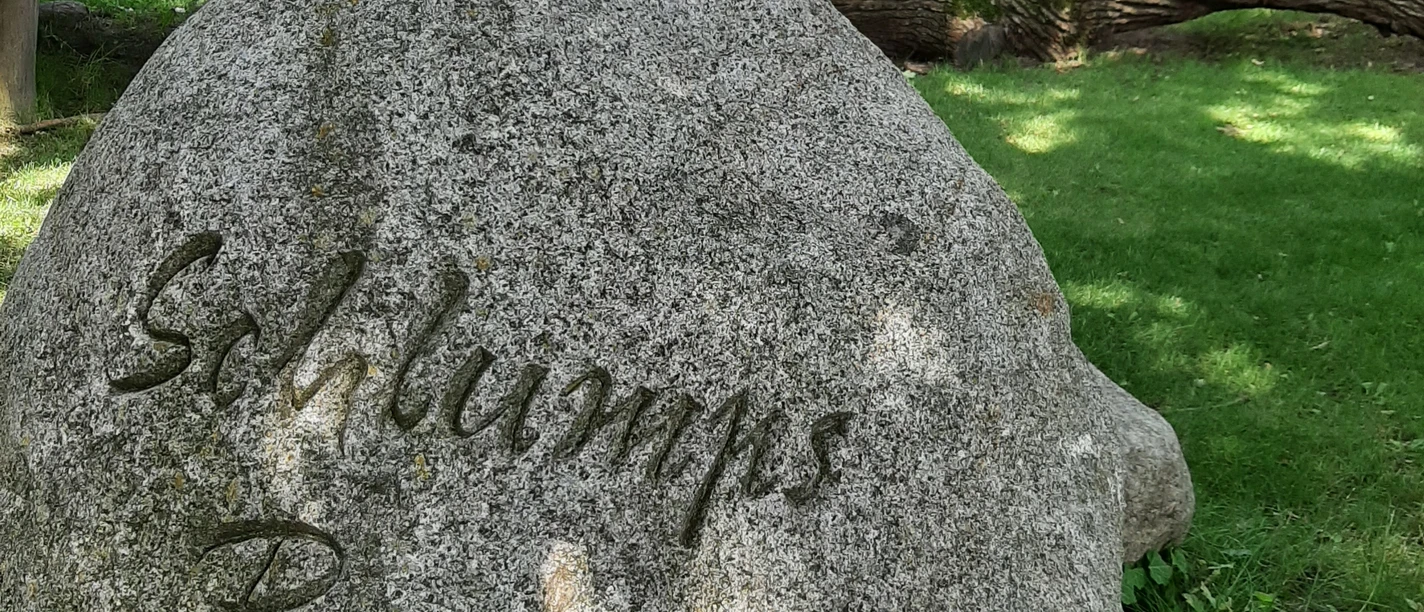 Großer Stein mit Inschrift „Schlumps Backs“, umgeben von Bäumen und einem traditionellen Fachwerkhaus.