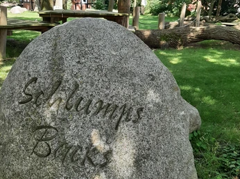 Schlumps Backs Großer Stein mit Inschrift „Schlumps Backs“, umgeben von Bäumen und einem traditionellen Fachwerkhaus.