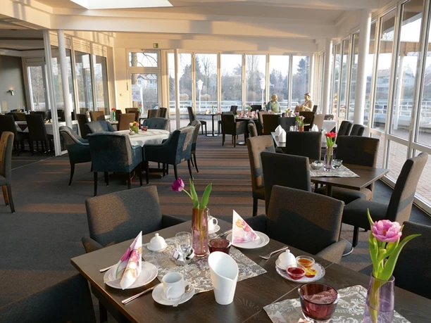 Helles, einladendes Restaurant mit gedeckten Tischen, Blumen und großen Fenstern mit Blick ins Freie.