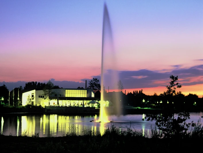 Ein großer Springbrunnen auf einem See im Vordergrund, beleuchtet bei Sonnenuntergang mit Gebäuden am Ufer.